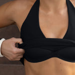 Black halter bikini top with deep plunge neckline, shown on model with hand adjusting fabric.